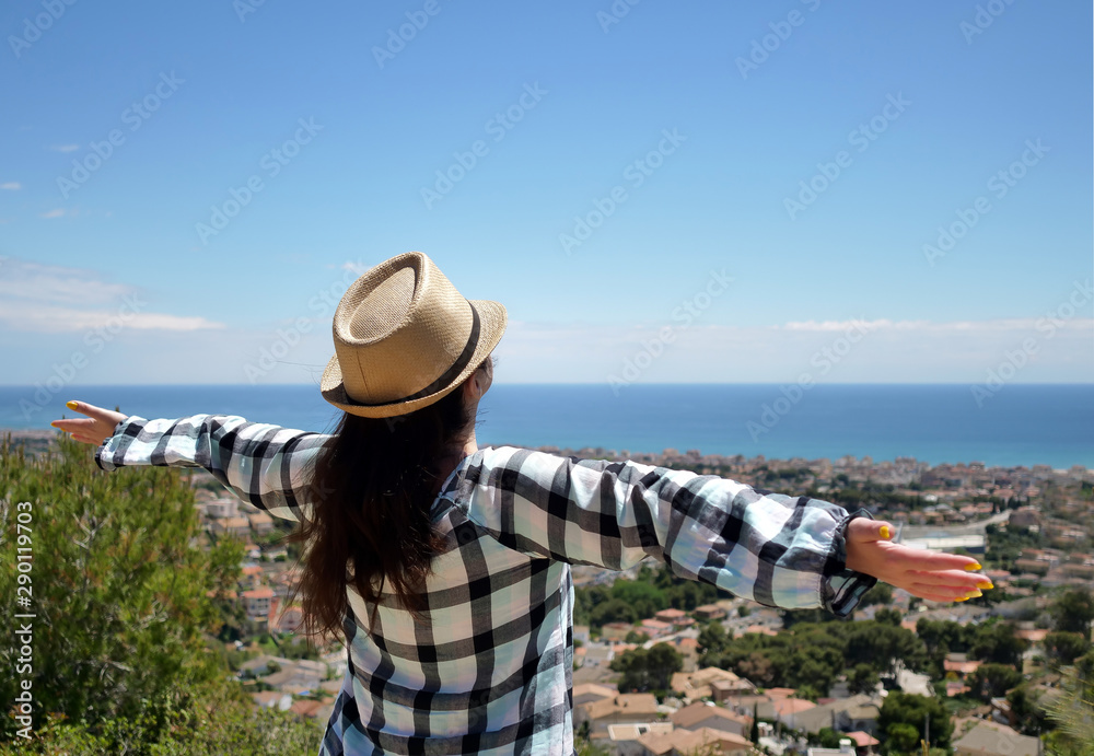 A young brunette girl in a hat stands on the mountain, spreading her arms wide apart, enjoying the journey