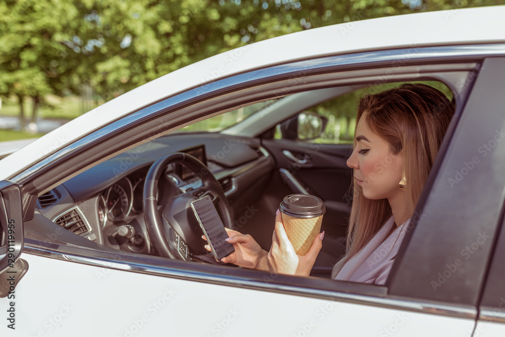 Naklejka premium girl driving car, stands in parking lot, summer waiting for friends and girlfriends, reads message on her smartphone, holds cup of tea her hands with coffee. Summer city background trees.