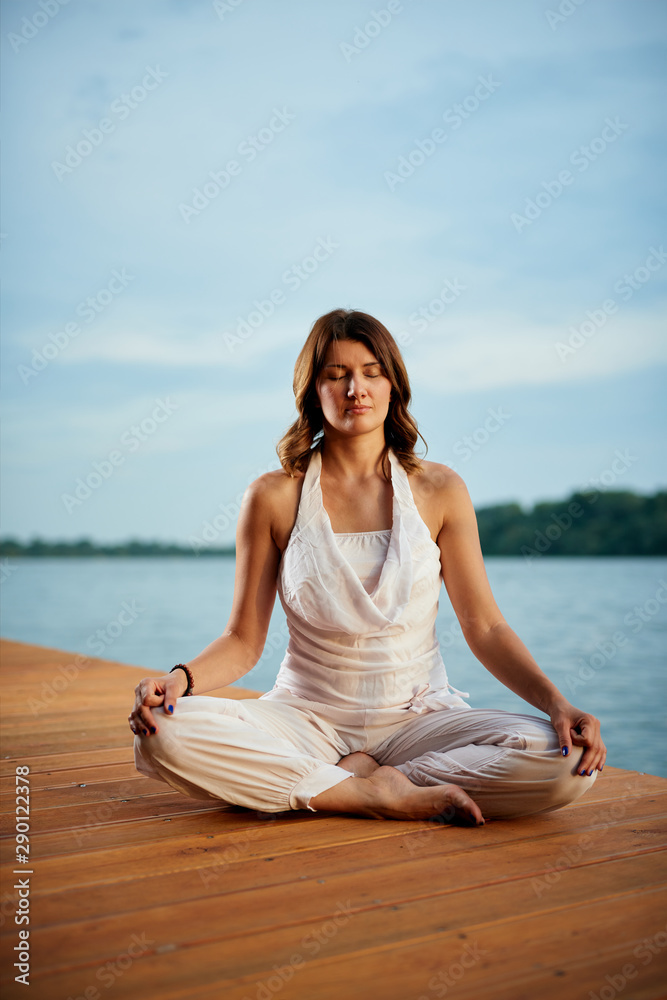 Beautiful caucasian brunette dressed in white outfit sitting near river and meditating. Yoga outdoor concept.