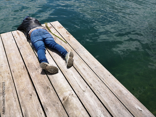 Junge in blauen Jeans und Lederjacke liegt auf den Brettern eines Bootssteg aus rustikalem Holz am Nesthauser See beim Lippesee im Sommer bei Sonnenschein in Paderborn in Ostwestfalen-Lippe photo