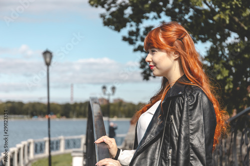 Cute happy young girl with red hair in a black leather jacket on a background of a city lake on a sunny day © Sergey