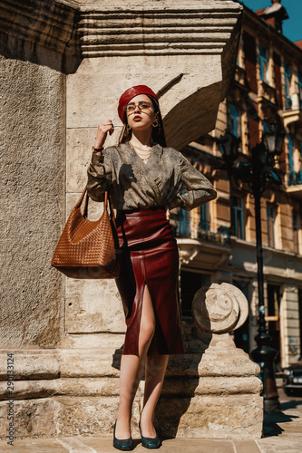 Outdoor full-length fashion portrait of elegant, luxury woman wearing trendy leather dark red pencil midi skirt, beret, snakeskin print blouse, shoes, sunglasses, holding brown bag, posing in street