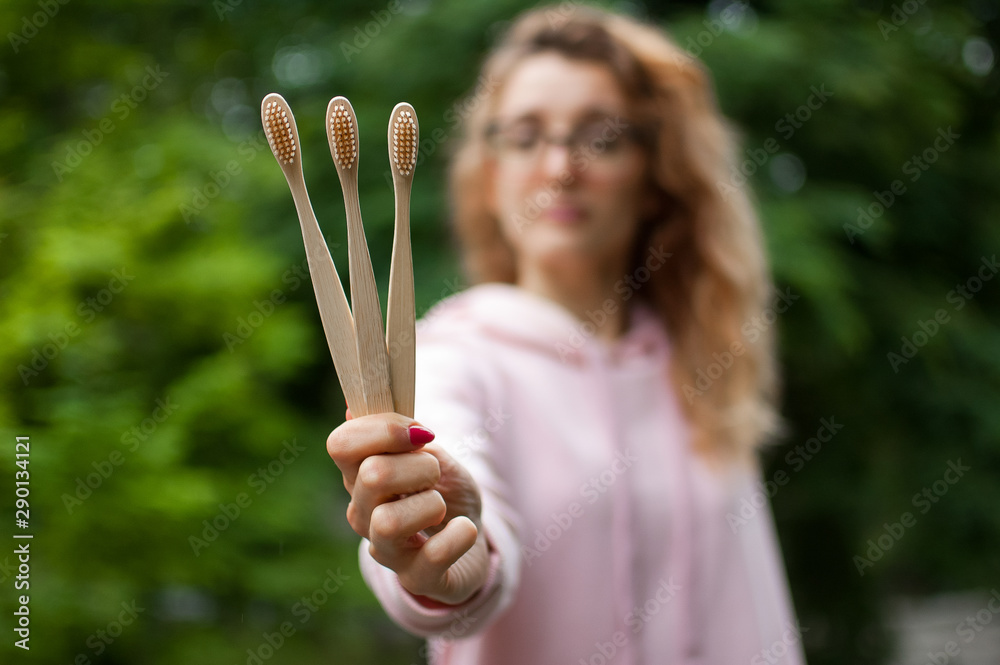 Outdoors portrait of young beautiful woman with blonde curly hair and eyeglasses holding three bamboo toothbrushes in hand. Green living concept Outdoors portrait of young beautiful woman with blonde curly hair and eyeglasses holding three bamboo toothbrushes in hand. Green living concept