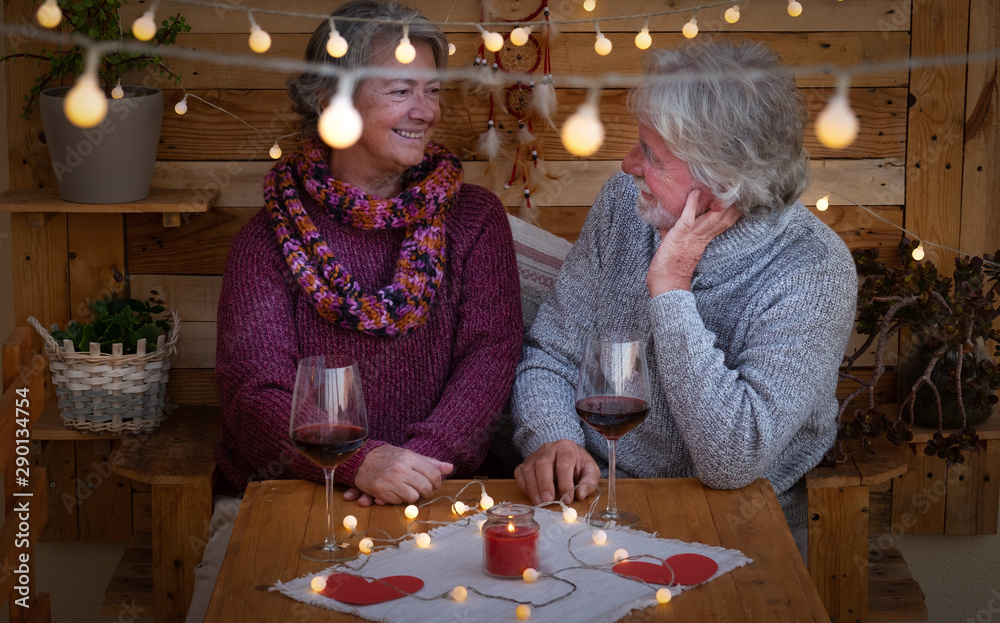 Fototapeta premium A smiling senior couple looking each other sitting outdoor in a romantic corner made by wood. Yellow lights. Two red wineglasses to celebrate a positive moment