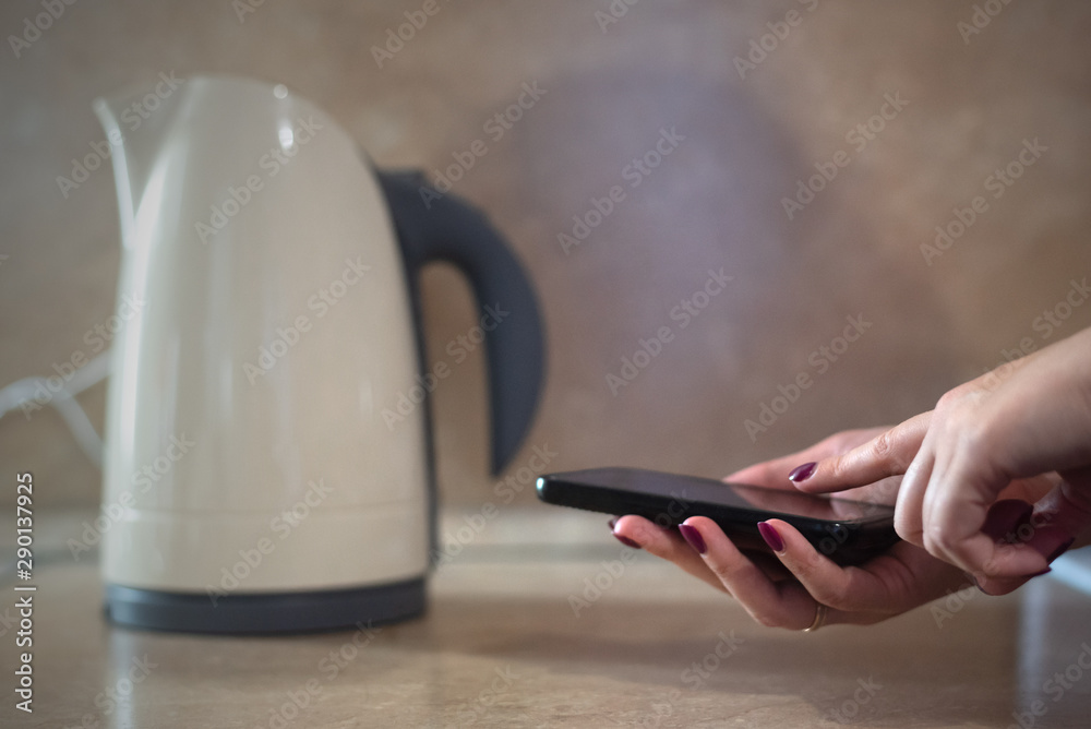 Fototapeta premium Woman is warming up a water in the kettle by a mobile phone with smart home application concept.
