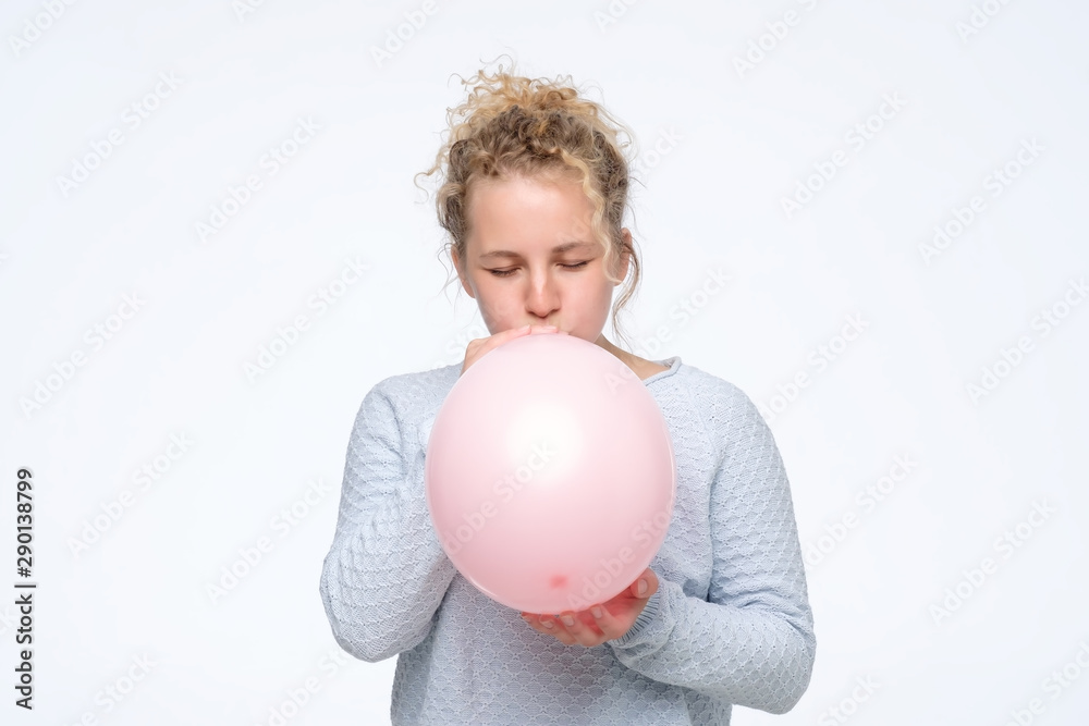 Young caucasian curly woman in gblue sweateris blowing pink balloon preparing for birthday party. Studio shot Young caucasian curly woman in gblue sweateris blowing pink balloon preparing for birthday party. Studio shot