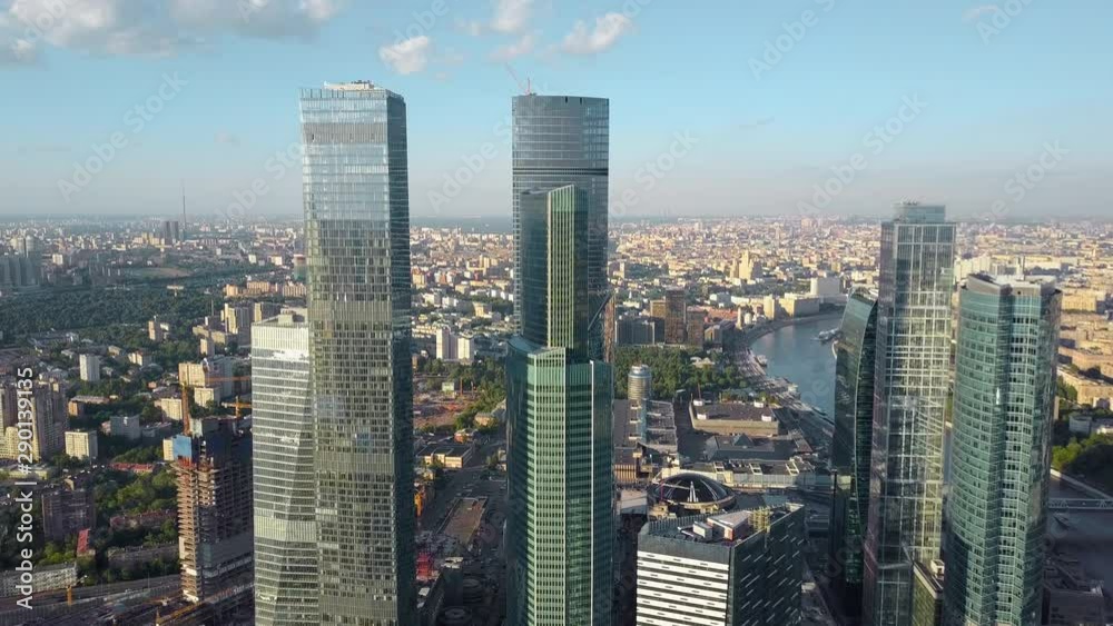 An aerial view of several shiny skyscrapers rising above the beautiful urbanscape. Multi storey buildings with wide roads between them, green recreational areas and winding river. Few clouds are An aerial view of several shiny skyscrapers rising above the beautiful urbanscape. Multi storey buildings with wide roads between them, green recreational areas and winding river. Few clouds are