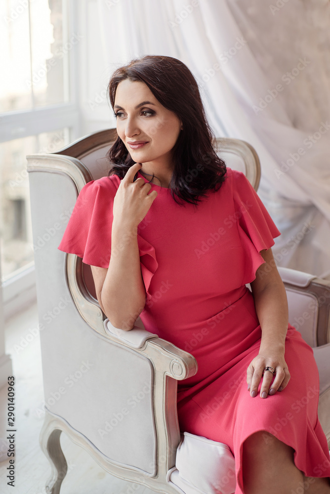 Beautiful middle-eged smiling woman model with dark hair wearing pink dress,posing near window in the classic room. Beautiful middle-eged smiling woman model with dark hair wearing pink dress,posing near window in the classic room.