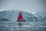 Beautiful red sailboat in the arctic next to a massive iceberg showing the scale. Cruising among floating icebergs in Disko Bay glacier during midnight sun season of polar summer Ilulissat, Disko Bay