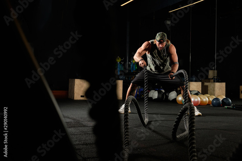 Muscular man in a sports uniform and a baseball cap performs an exercise with ropes in the gym
