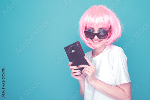 90s style concept. Young woman with pink hail holding glasses and cassette standing in the blue colored studio