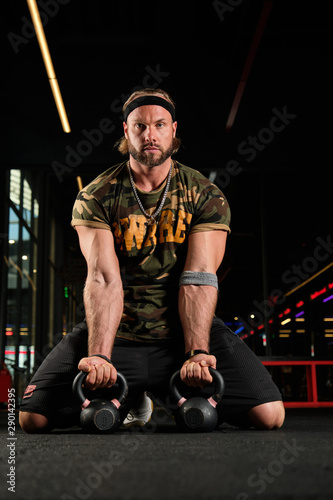 Muscular man in a sports uniform is kneeling in the gym holding weights in his hands and looking at the camera