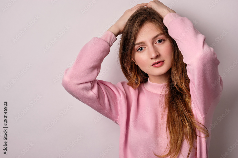 A blonde girl in a pink sweatshirt stands on a white background and looks into the frame with her hands on her head