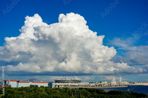 A huge cloud over the city. Park near the river. Needles of the stadium support the cloud.