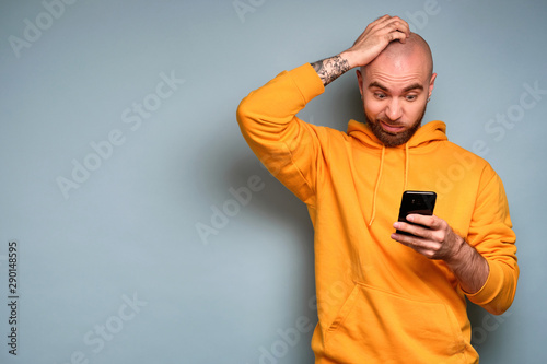 A bearded guy in a yellow sweatshirt stands on a blue background and looks in horror at the smartphone with his hand on his head © bestsenny