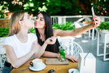 Two charming smiling woman taking selfie on smartphone, sitting at cafeteria