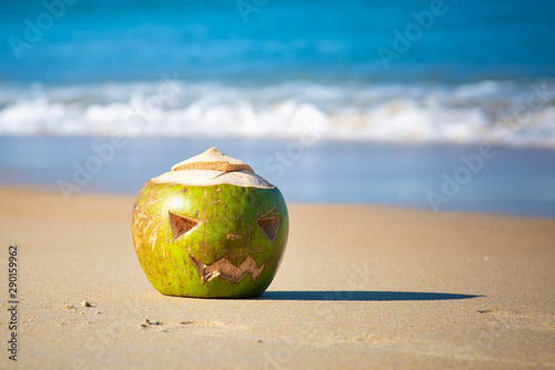 Green coconut with a face carved on it, like a halloween pumpkin. Lies in the sand on a tropical beach on a background of waves. Holiday trip concept.