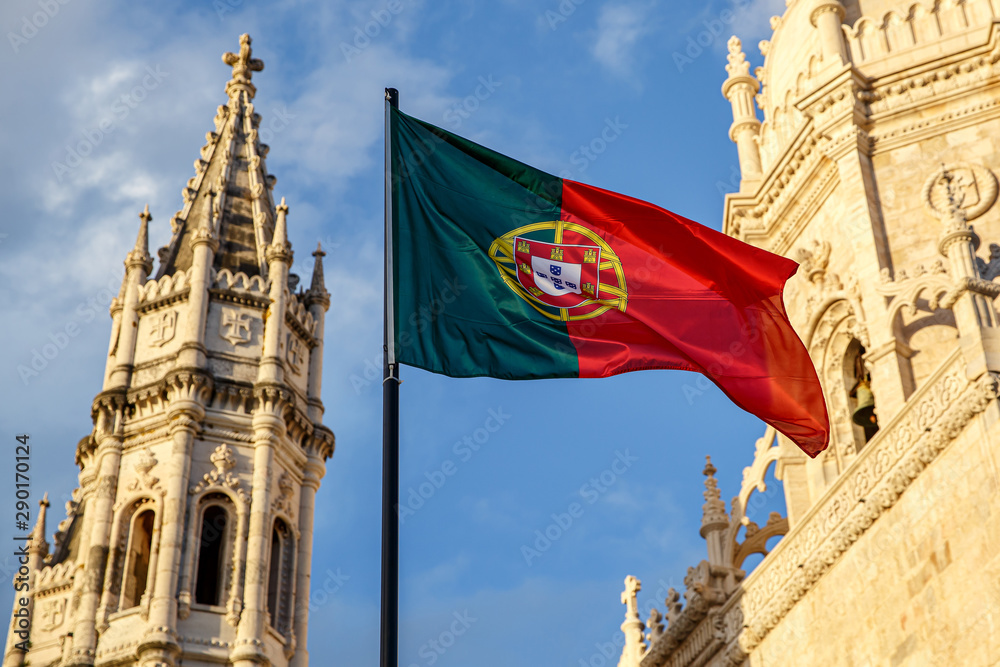 Portuguese flag waving in front of a blue sky and monastery.