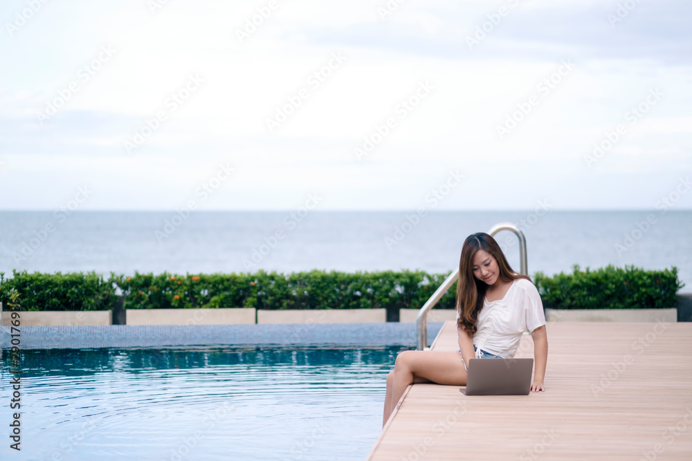 A beautiful asian woman using and typing on laptop computer while sitting by swimming pool
