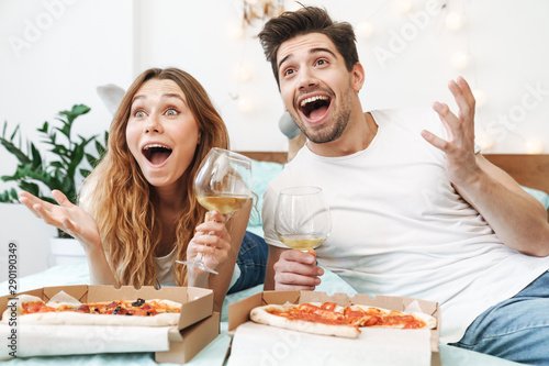 Image of joyful couple drinking wine and eating pizza on bed at home