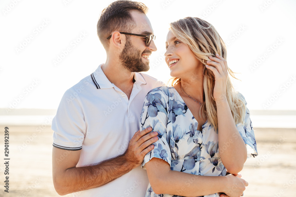 Photo of beautiful happy couple smiling each other and hugging while walking on sunny beach Photo of beautiful happy couple smiling each other and hugging while walking on sunny beach