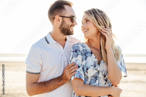 Photo of beautiful happy couple smiling each other and hugging while walking on sunny beach