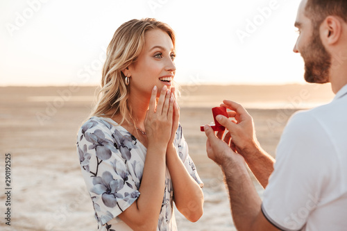 Photo of romantic man making proposal to his amazed woman with ring while walking on sunny beach photo