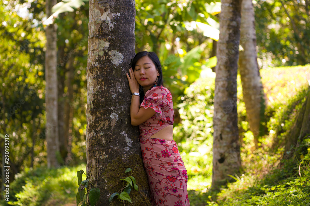 young beautiful and relaxed Asian Korean woman in elegant summer dress enjoying serene the beauty of nature leaning dreamy on tree trunk at fresh tropical rainforest