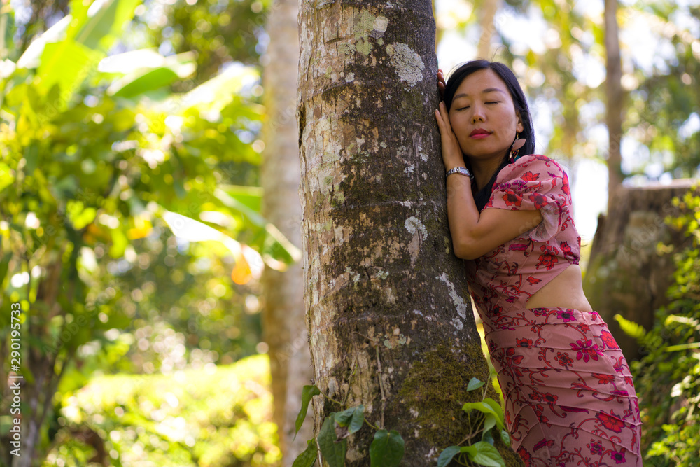 young beautiful and relaxed Asian Korean woman in elegant summer dress enjoying serene the beauty of nature leaning dreamy on tree trunk at fresh tropical rainforest young beautiful and relaxed Asian Korean woman in elegant summer dress enjoying serene the beauty of nature leaning dreamy on tree trunk at fresh tropical rainforest