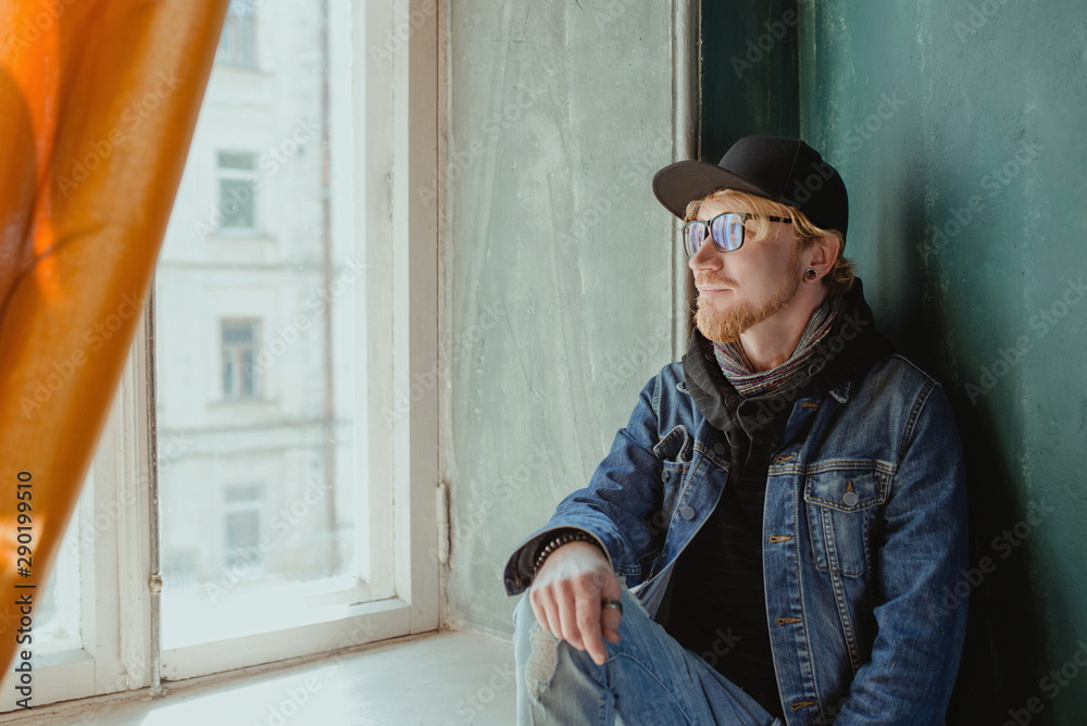 portrait of modern redhead handsome stylish man in black hat and Jean jacket and glasses alone by the window. Fashion concept portrait of modern redhead handsome stylish man in black hat and Jean jacket and glasses alone by the window. Fashion concept
