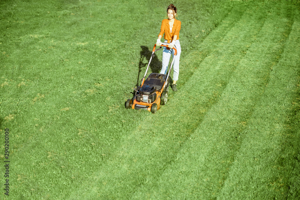 Beautiful young woman cutting grass with gasoline lawn mower, gardening on the backyard in the countryside, view from above Beautiful young woman cutting grass with gasoline lawn mower, gardening on the backyard in the countryside, view from above