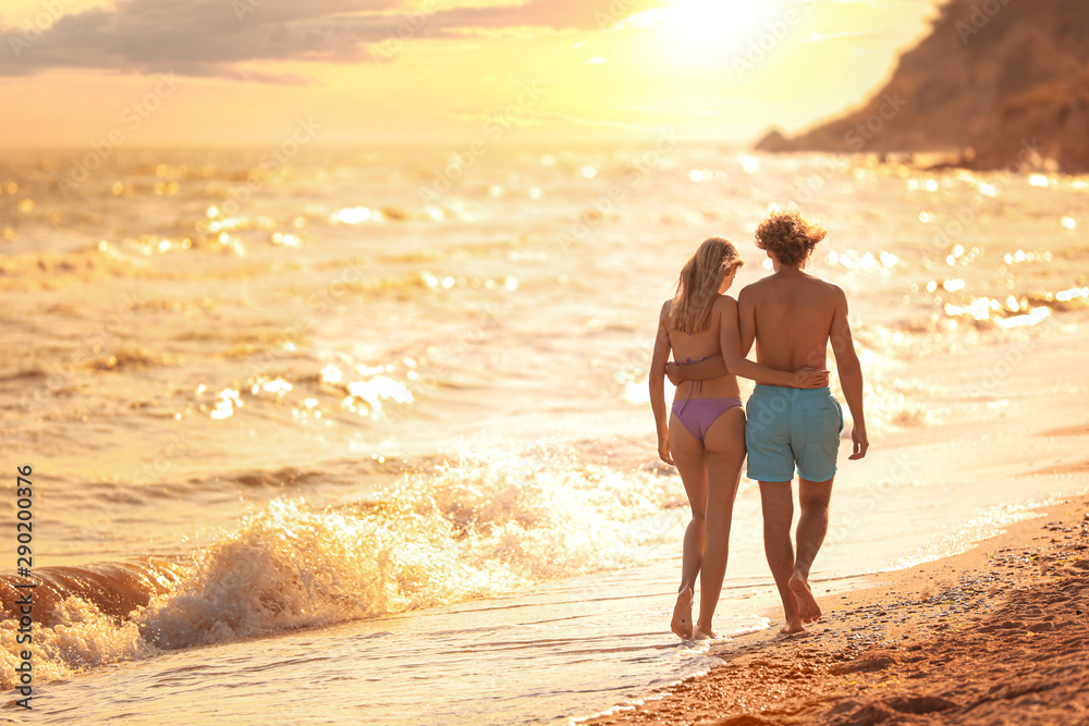 Young woman in bikini and her boyfriend walking on beach at sunset. Lovely couple - obrazy, fototapety, plakaty 