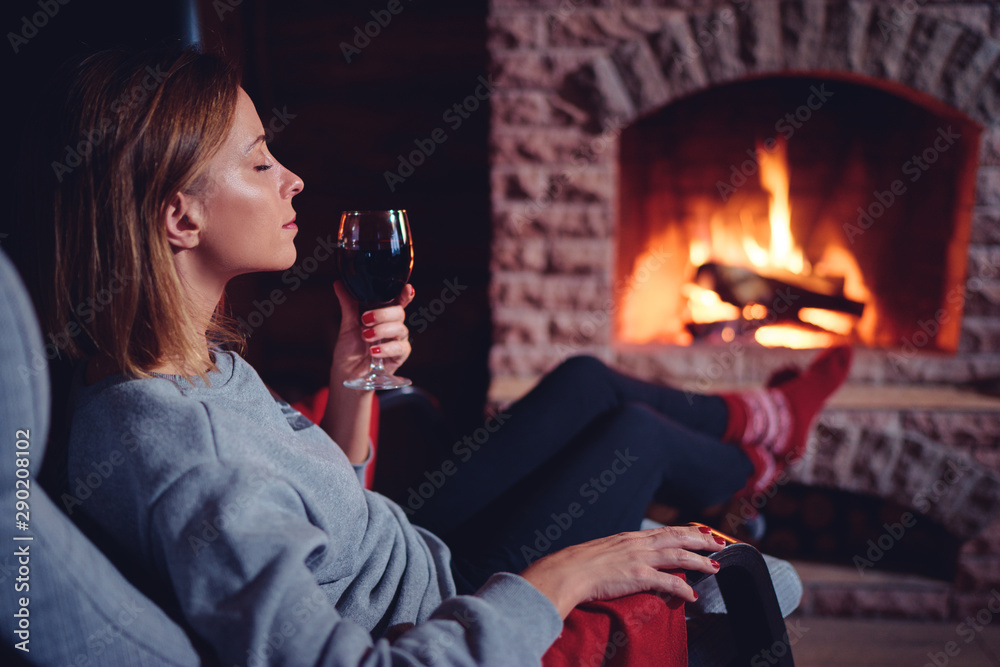 Cozy home. Close up of young woman drinking red wine near the fireplace. - obrazy, fototapety, plakaty 