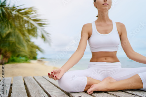 Yoga and meditation. Close up of young woman in lotus pose on wooden deck with sea view.