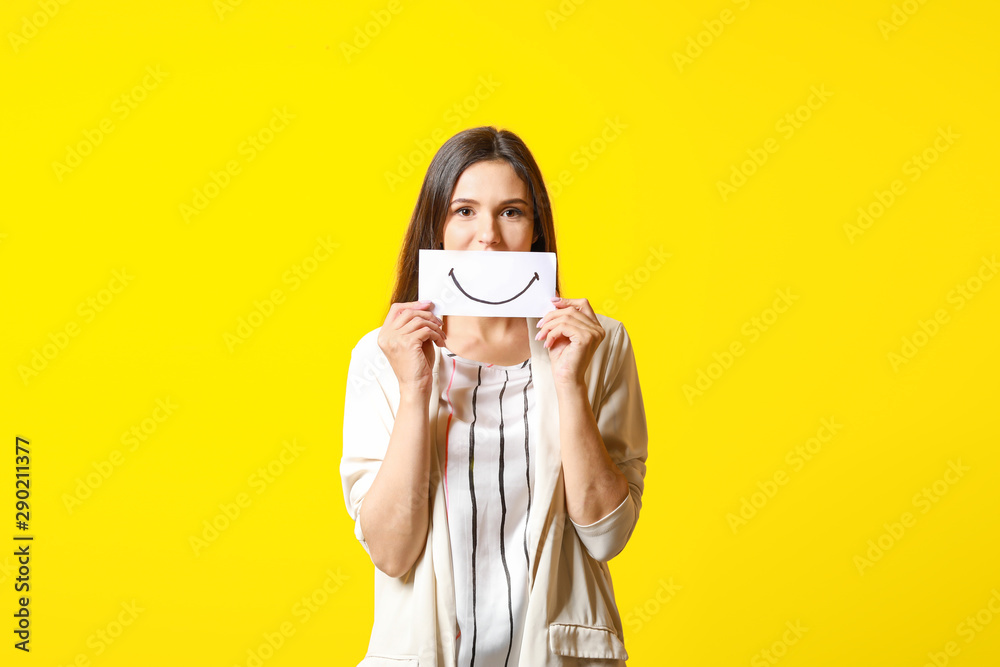 Woman hiding mouth behind sheet of paper with drawn smile on color background Woman hiding mouth behind sheet of paper with drawn smile on color background