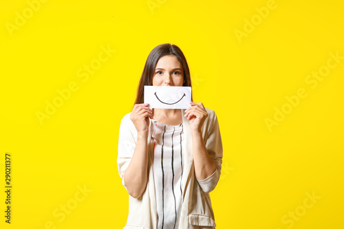 Woman hiding mouth behind sheet of paper with drawn smile on color background