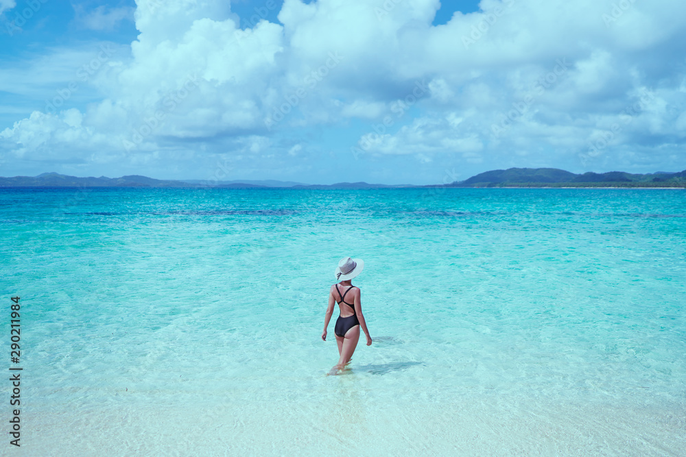 Vacation on the seashore. Back view of young woman in hat bathing on the beautiful tropical white sand beach.