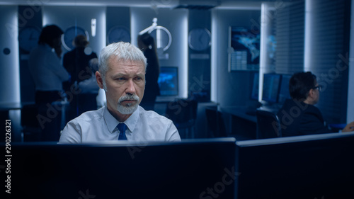 Portrait of the Concerned Senior Expert Working on Computer in the Monitoring Control Room with Teams Solving Problems in the Background. photo
