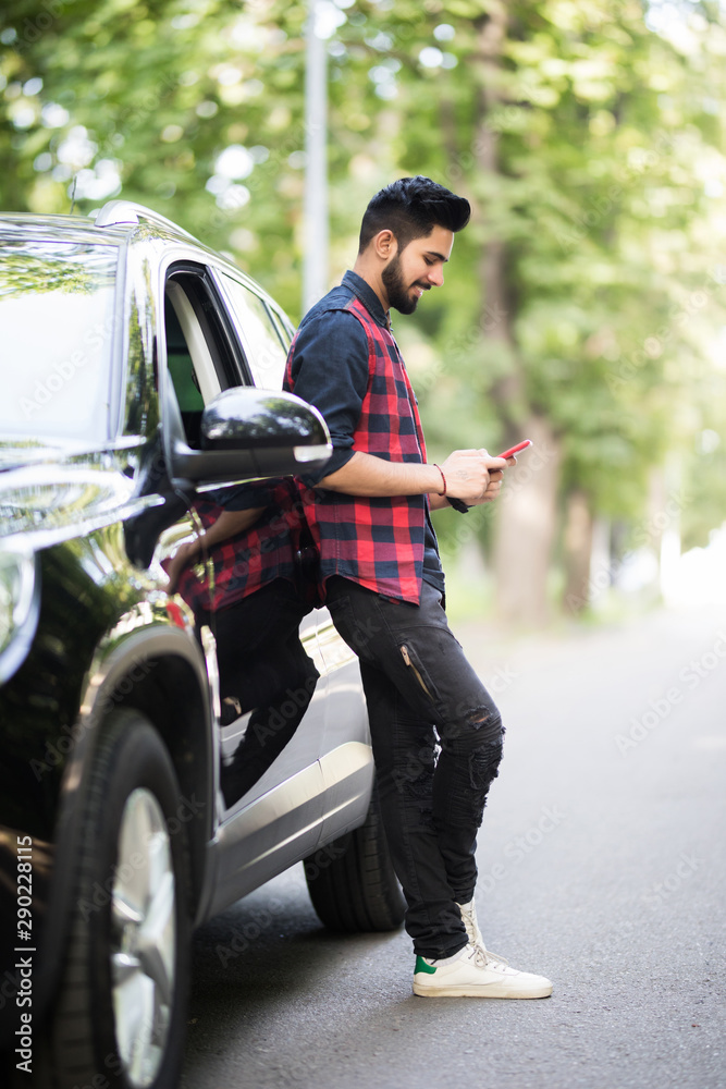 Inviting friend for a journey. Handsome young man use the mobile phone and smiling while leaning at his car Inviting friend for a journey. Handsome young man use the mobile phone and smiling while leaning at his car