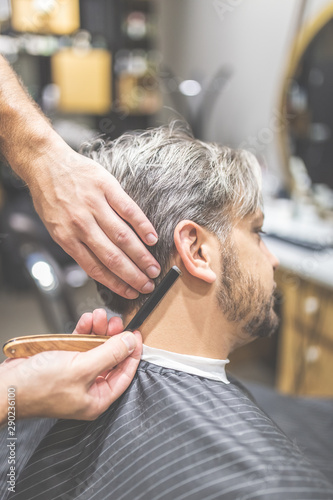 Making hair look magical. Side view of young bearded man getting shaved with straight edge razor by hairdresser at barbershop