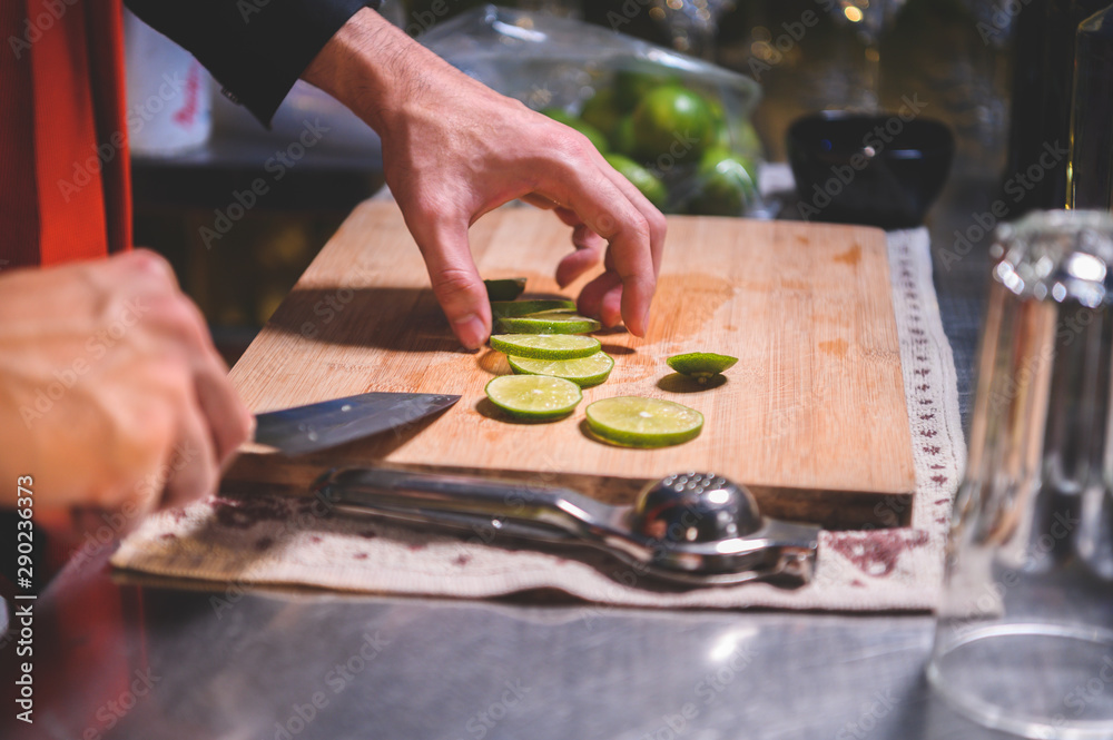 Closeup of professional bartender hand slicing lemon for making lemonade juice by knife in night club. Chef making drinks for guest in pub restaurant. Food and beverage concept.