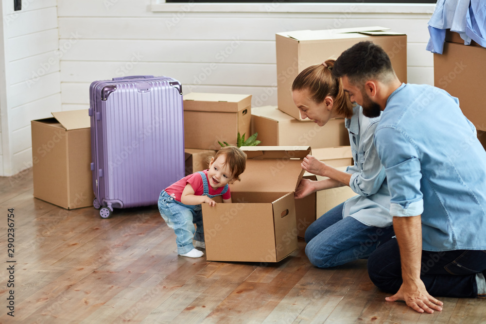 Couple wear blue shirt peek box which carries kid wear denim overalls. Background moving boxes, suit and flower in pot. Family smile Couple wear blue shirt peek box which carries kid wear denim overalls. Background moving boxes, suit and flower in pot. Family smile