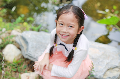Happy little child girl sitting near the pond in the nature garden.