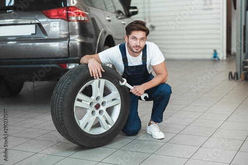 young handsome mechanic wearing uniform working in car service department fixing flat tire looks pleased