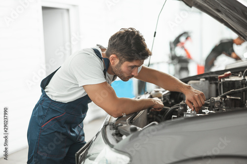 young handsome smiling mechanic in uniform fixing motor in car bonnet working in service department