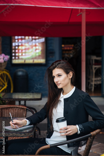 Young beautiful brunette girl, office clothing style, sitting on the street at a table in a cafe, street model posing style, makes notes in a notebook, talking on the phone