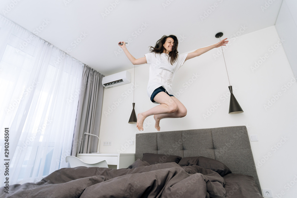 Young adult woman waking up and jumping on bed with smartphone - obrazy, fototapety, plakaty 