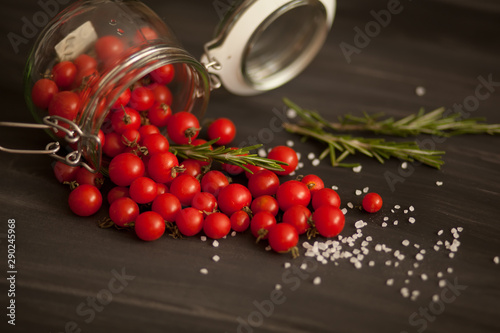 red small cherry tomatoes summer and autumn harvest in a glass jar with salt pepper rosemary on a black background scattered