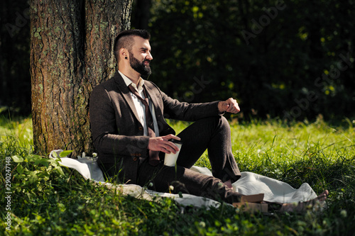 Young attractive bearded business man sitting on green grass under tree and resting in park. Read book, drink coffee. Relaxed, tired of work, lunch break.