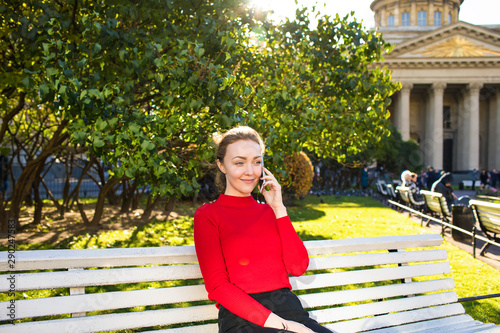 Young stylish hipster girl talking via smartphone gadget while sitting outdoors on bench during free time in vacation. Female traveler having mobile phone conversation while relaxing in park #290247583